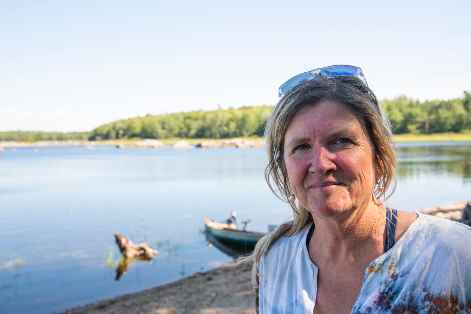 Woman with sunglasses on her head standing at a rocky lakeshore beside a canoe