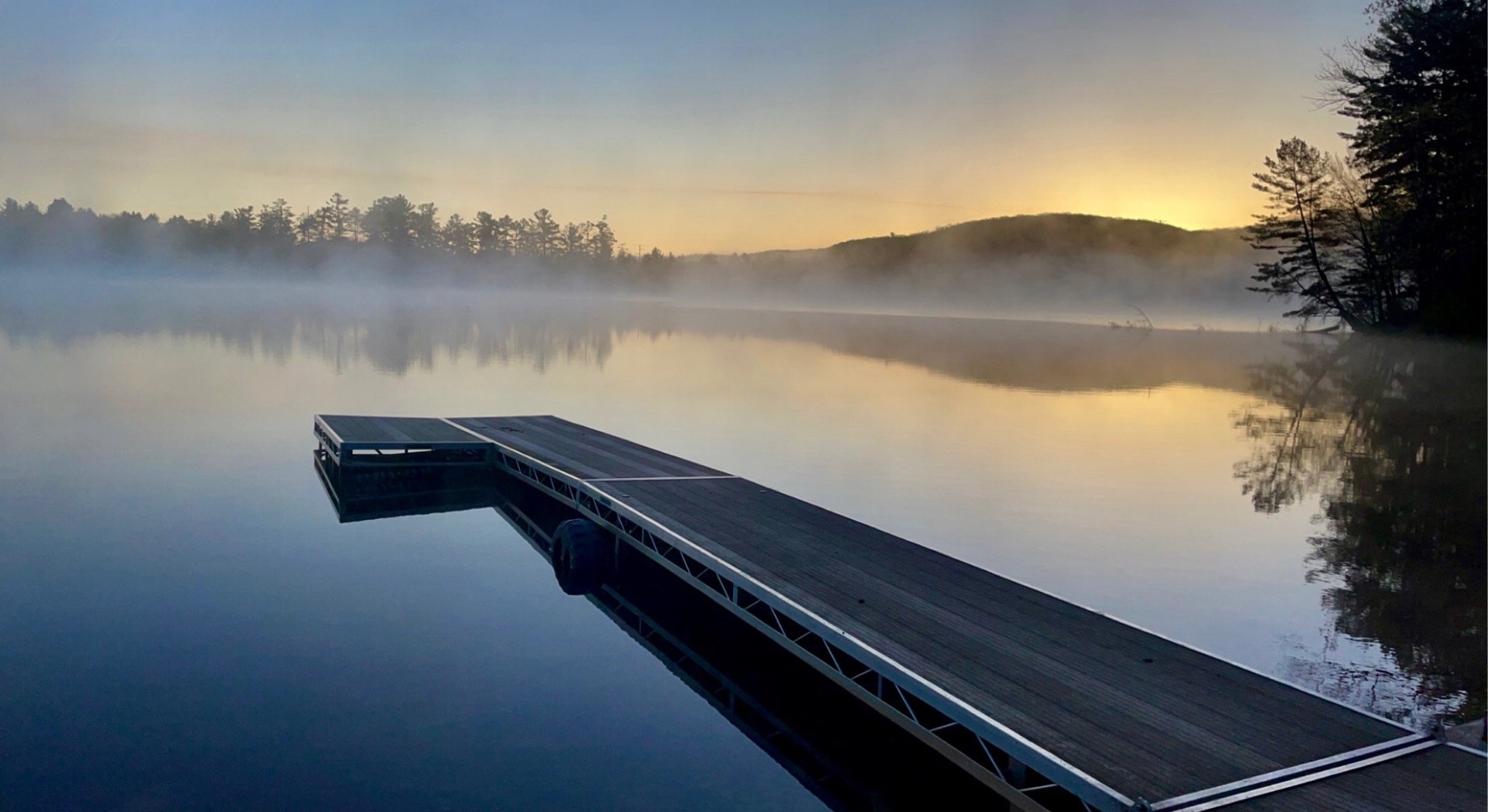 A dock reaching into a still northern lake at dawn, mist over the water and sunrise behind the treeline