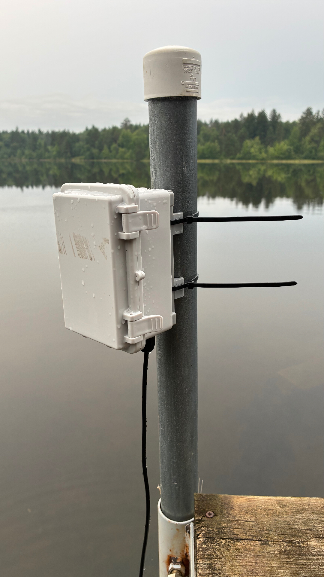 A weather sensor enclosure mounted on a dock post above a calm lake, with forested shoreline in the background