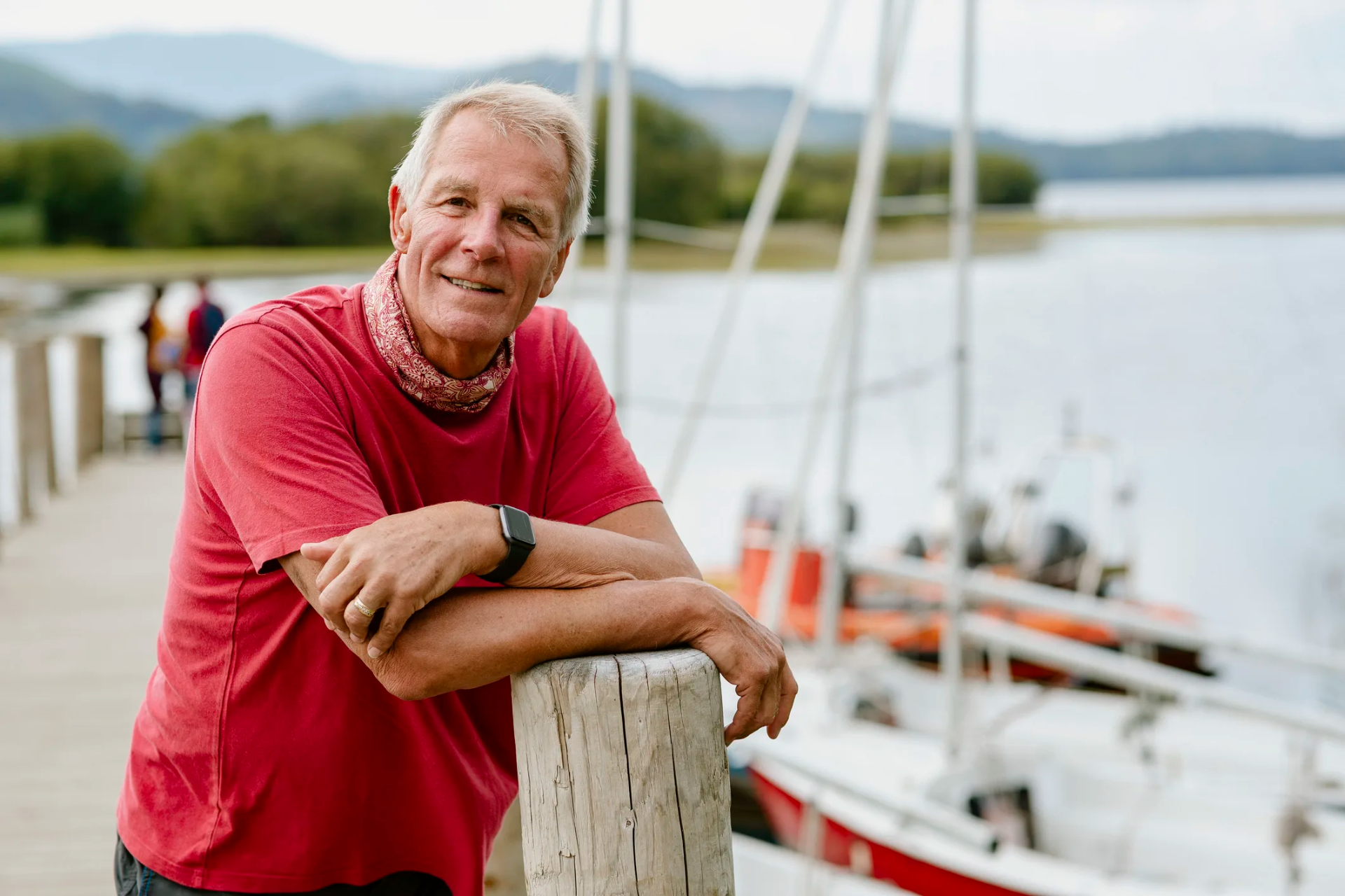 Man in a red shirt leaning on a dock post at a lakeside marina, sailboats and hills behind him
