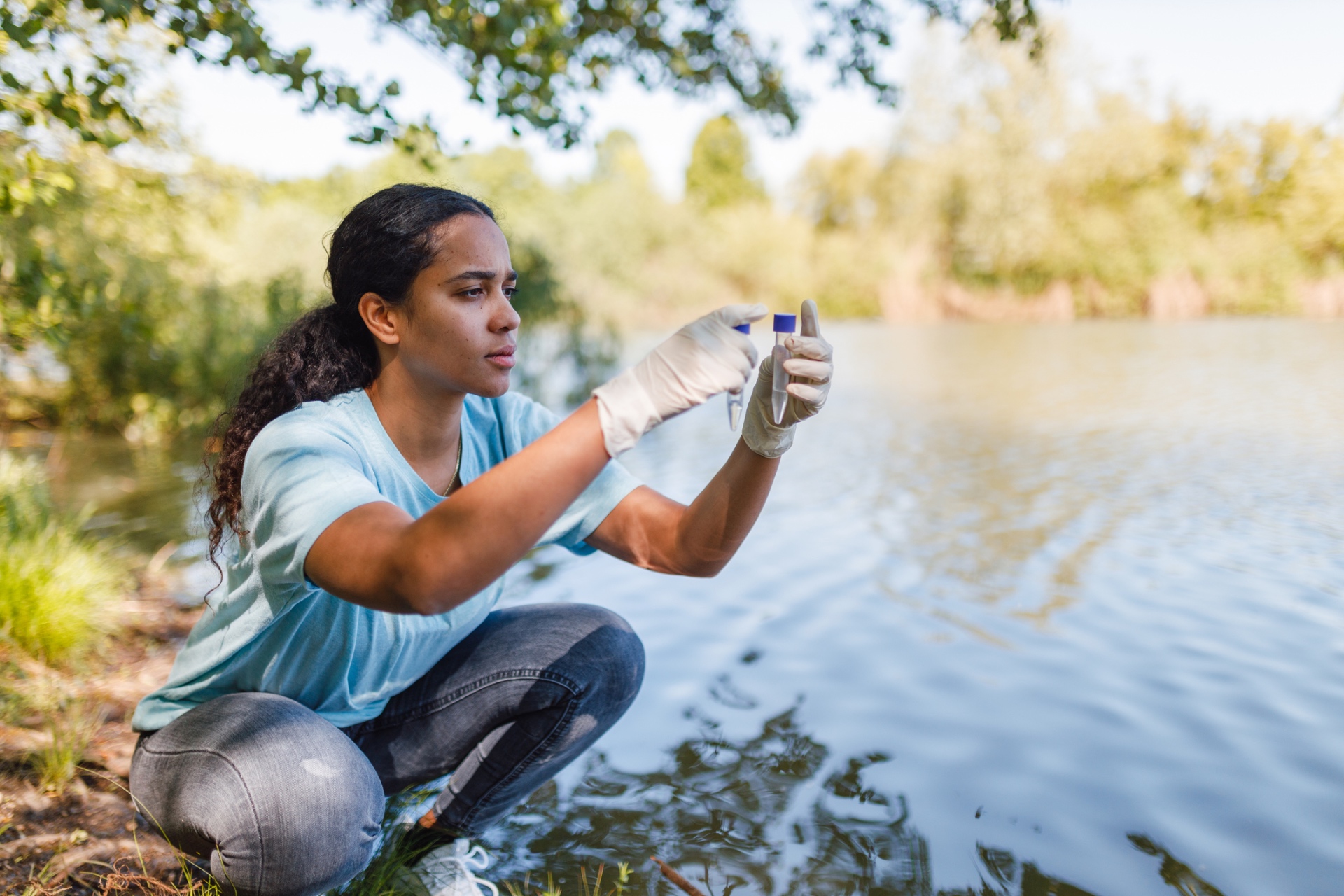 A volunteer kneeling at the shoreline holding a water sample vial up to the light