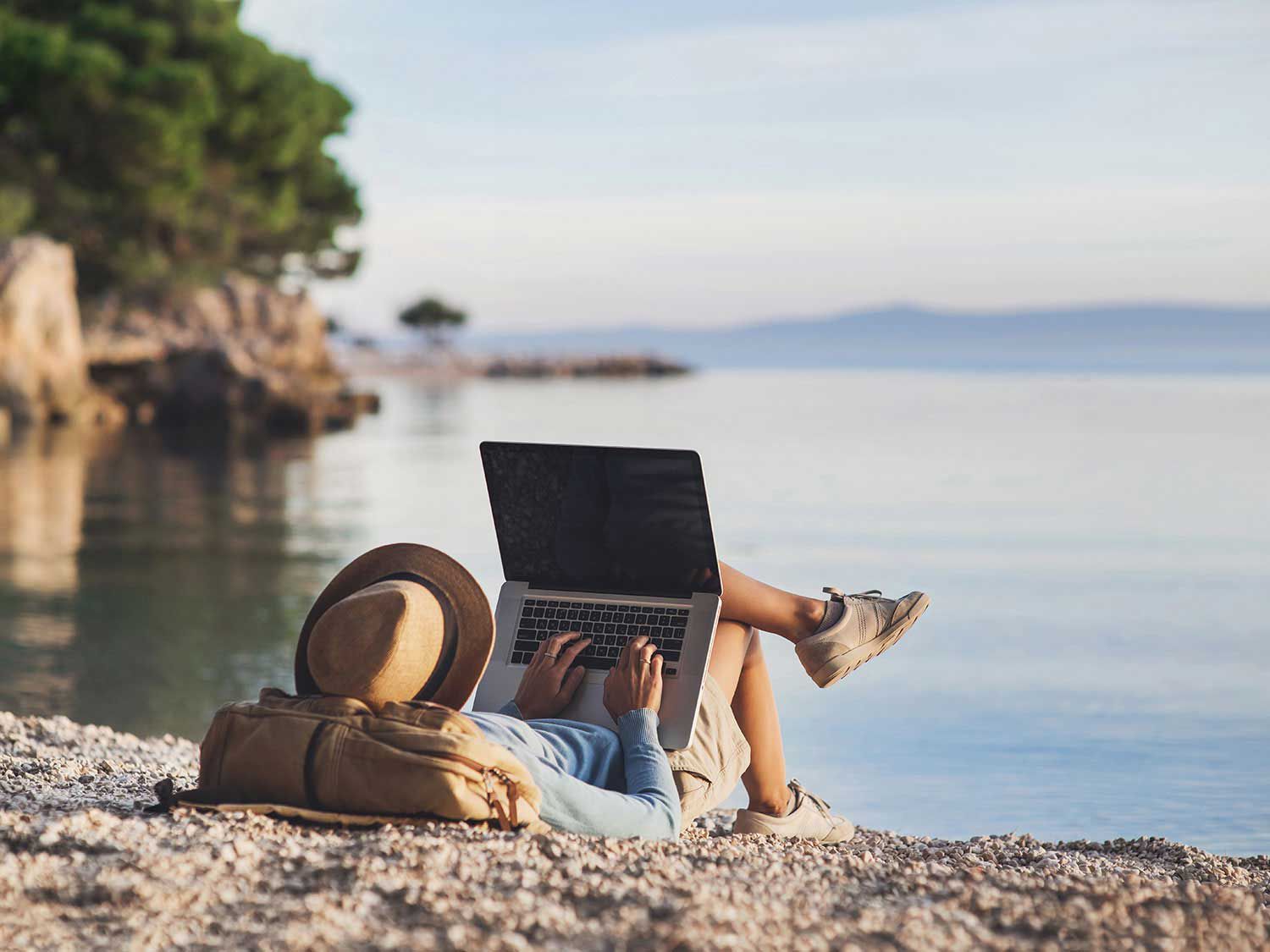 A person relaxing on a rocky shoreline with a laptop, looking out at calm water