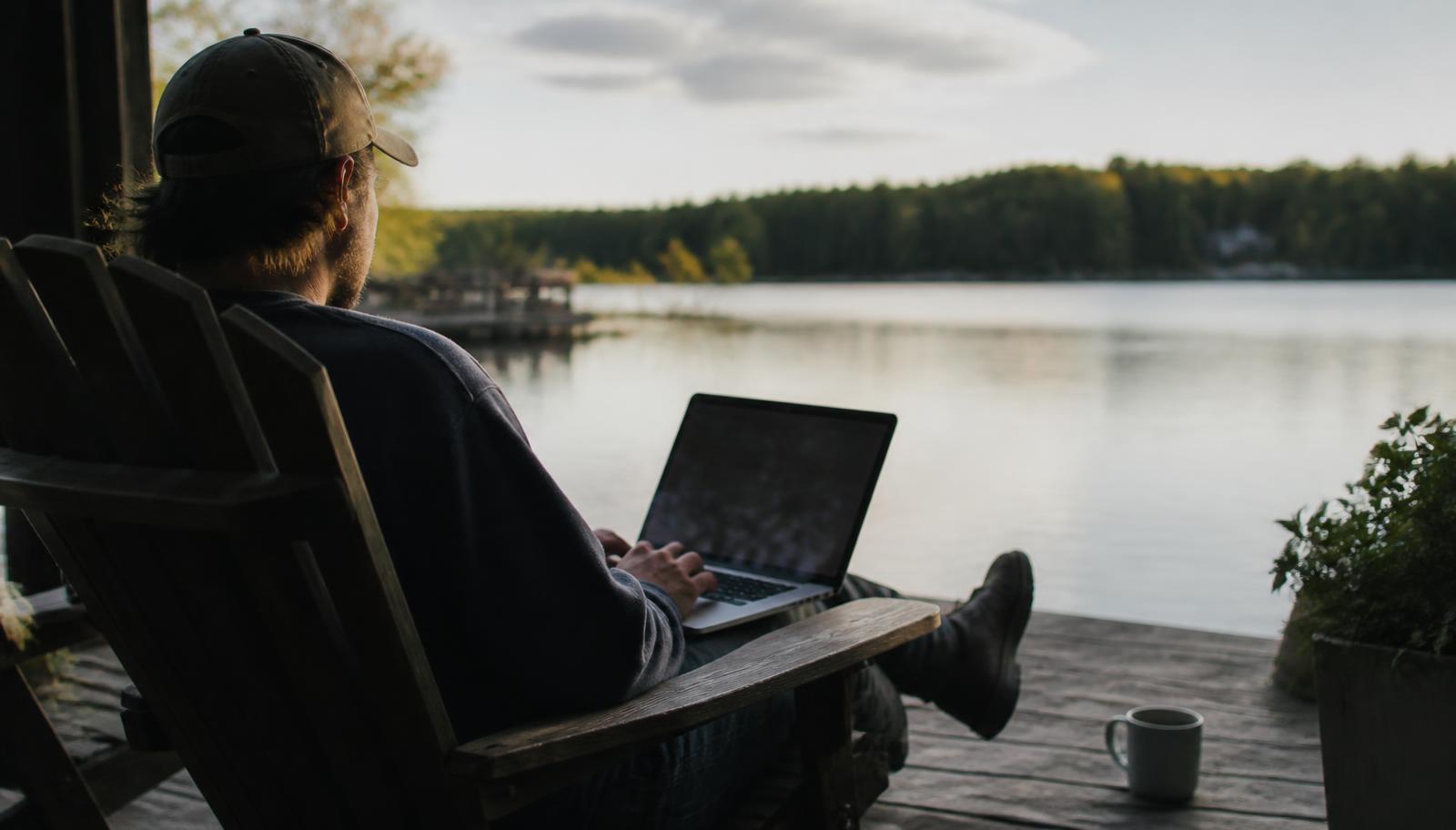 A man sitting on a lakeside deck with a laptop, calm water and treeline behind them